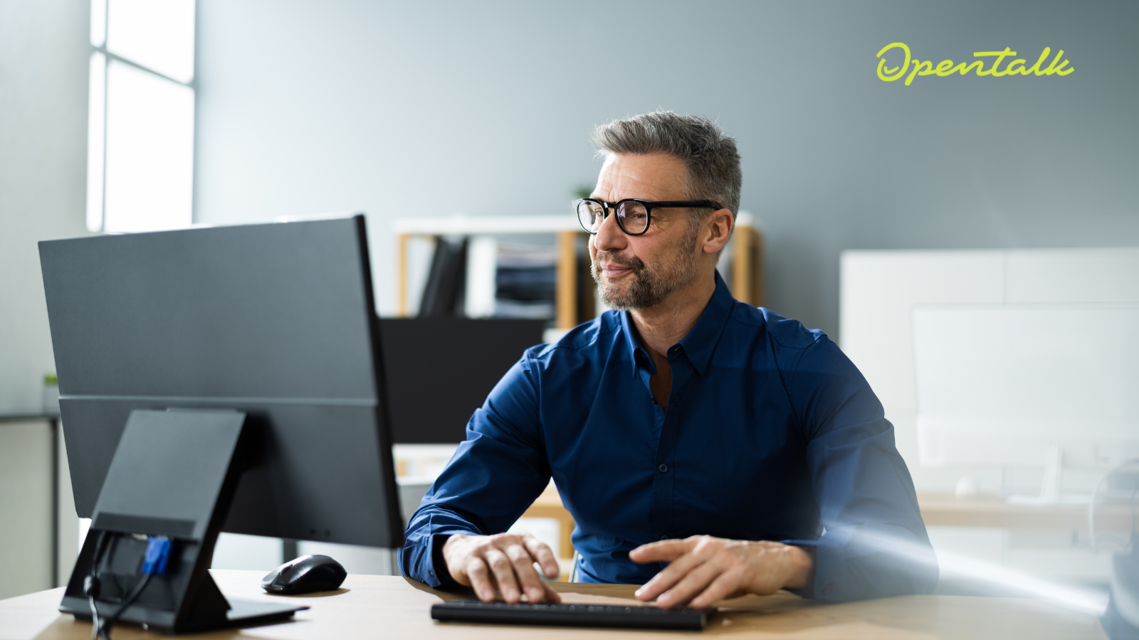Ein Mann sitzt an einem modernen Arbeitsplatz vor einem Computer-Monitor. In der oberen rechten Ecke des Bildes steht in gelber Schrift "OpenTalk".