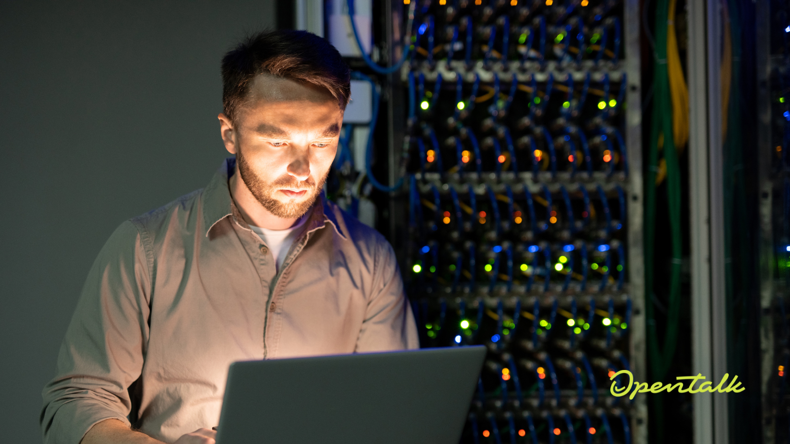 Ein Techniker im Serverraum prüft Daten auf einem Laptop, während neben ihm Server-Racks mit leuchtenden Status-LEDs stehen. In der unteren rechten Ecke ist das OpenTalk-Logo eingeblendet.