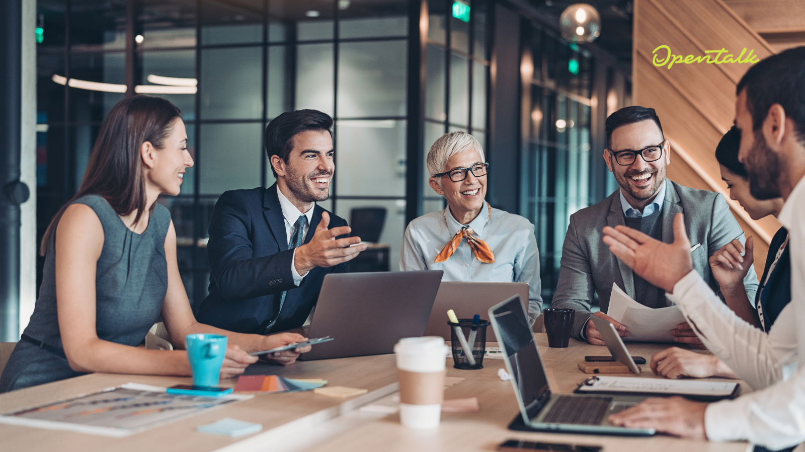 	Mehrere Geschäftsleute sitzen an einem Konferenztisch, unterhalten sich lachend und arbeiten mit Laptops, Dokumenten und Kaffeebechern in einem modernen Büro.
