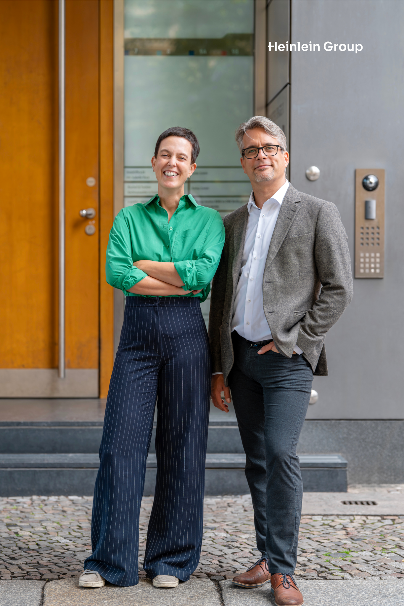 Jutta Horstmann und Peer Heinlein stehen nebeneinander vor einem Hauseingang. Rechts ist das Logo ‚Heinlein Group‘ sichtbar.