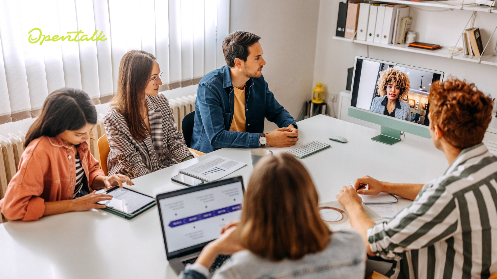 Fünf Personen sitzen in einem Konferenzraum an einem Tisch und nehmen an einem Videomeeting teil. Auf einem Bildschirm ist eine Frau zu sehen, die virtuell zugeschaltet ist. Auf dem Tisch liegen Laptops, Tablets und Unterlagen. Links oben im Bild ist das OpenTalk-Logo eingeblendet.