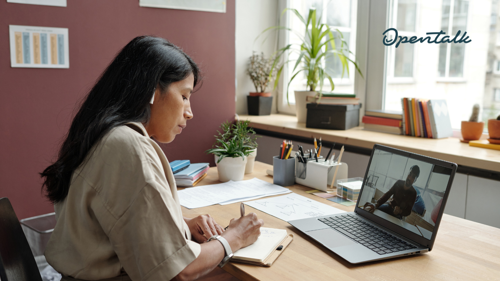 Eine Frau sitzt an einem Schreibtisch in einem hellen Büro und macht sich Notizen in einem Notizbuch, während sie über einen Laptop an einer Videokonferenz teilnimmt. Oben rechts im Bild ist das OpenTalk-Logo eingeblendet.